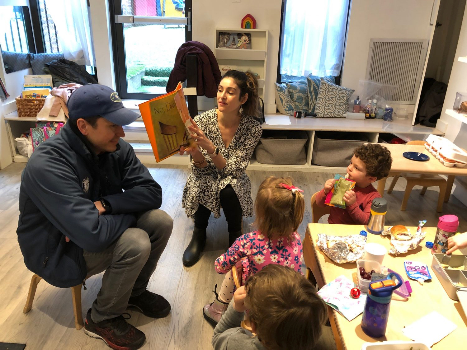 a mother and father reading to a classrooom of children as they eat